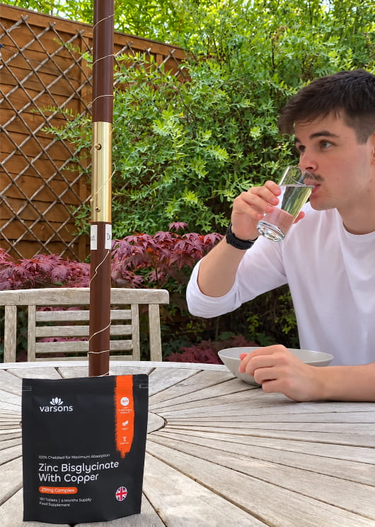 Customer drinking a glass of water at a garden table next to a Varsons Zinc Bisglycinate with Copper pouch